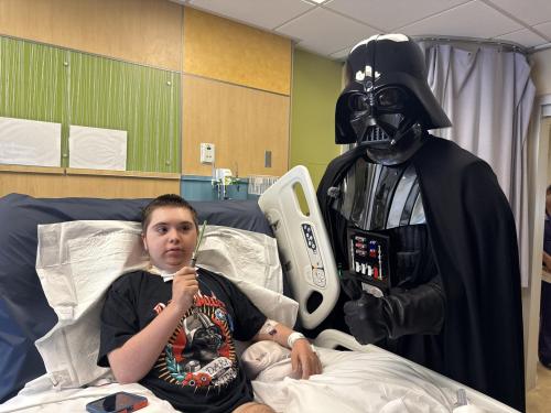 A special bedside visit brought a memorable moment to the Pediatric Intensive Care Unit at Miller Children’s & Women’s Hospital, where Michael, a smiling 14 year old patient, showed off his lightsaber and Darth Vader T shirt alongside the iconic character.