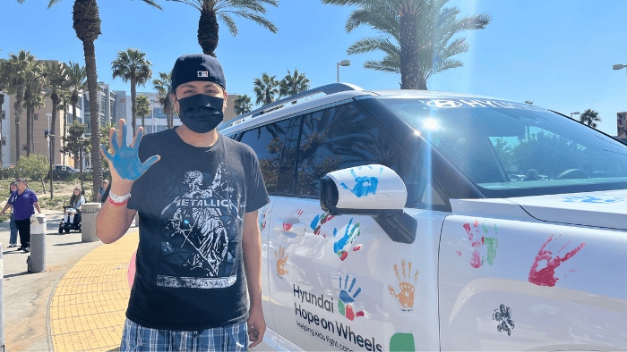 Boy holds hand up with colorful handprint next to painted Hyundai car.