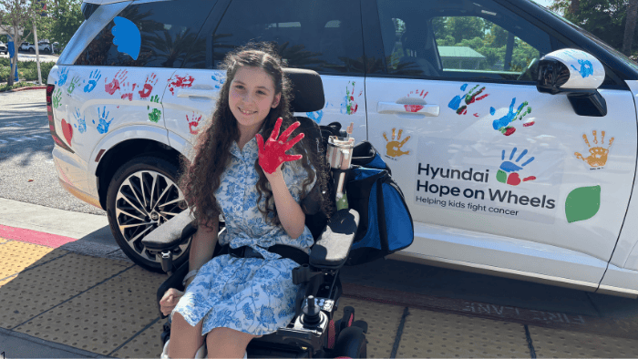 Abigail, a 16-year-old patient at Miller Children’s & Women’s Hospital, stands in front of a newly painted Hyundai vehicle, proudly holding up her red-painted hand after participating in the Hyundai Hope on Wheels Handprint Ceremony to honor Childhood Cancer and Sickle Cell Awareness Month.