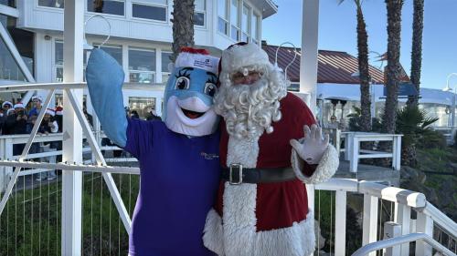 Santa makes a grand coastal arrival at Parker’s Lighthouse, stepping off his yacht to greet Millie the Dolphin, mascot of Miller Children’s & Women’s Hospital, while holiday excitement builds with eager children waiting nearby.