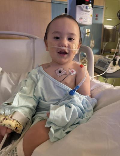 Makoa pictured in patient room during recovery, wearing a light blue gown and connected to monitoring equipment as part of his specialized care.