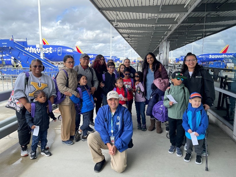 Stramski Children's Developmental Center patients and their caregivers stand close together for a photo outside a boarding gate at the Long Beach Airport. The scene behind them includes three big, blue Southwest branded airplanes.